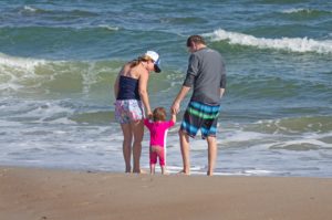 A mom and dad hold their toddler daughter by the arms, while facing the beach during their family vacation in Emerald Isle, NC.